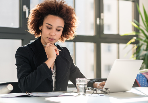 Woman in business attire sitting at a desk, working on a laptop with documents and a glass of water nearby in a brightly lit office.
