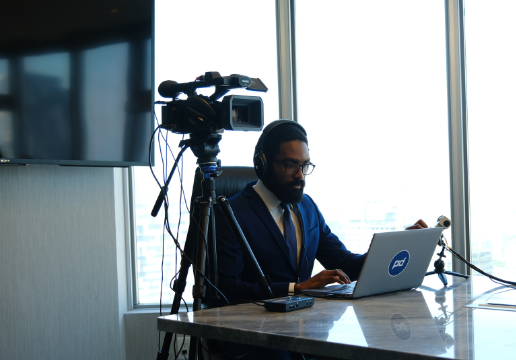 A man in a suit sits at a desk with a laptop, wearing headphones and speaking into a microphone, with a video camera set up nearby in an office setting.