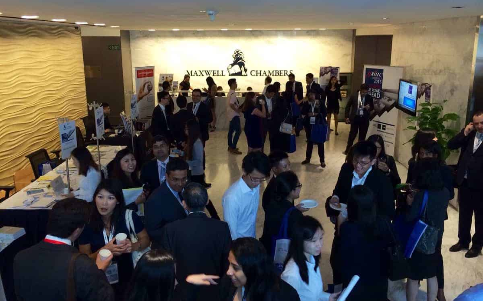 A group of people in business attire network and converse in the lobby of Maxwell Chambers, surrounded by informational booths and banners.