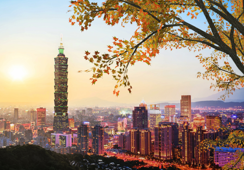Cityscape of Taipei at sunset, featuring Taipei 101 skyscraper, illuminated buildings, and autumn leaves in the foreground.