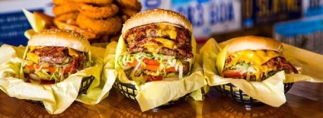 Three cheeseburgers with lettuce, tomato, and cheese in baskets lined with yellow paper, with a stack of onion rings in the background.