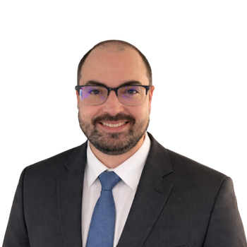 A man with short dark hair, beard, and glasses wears a dark suit, white shirt, and blue tie, smiling at the camera against a plain light background.