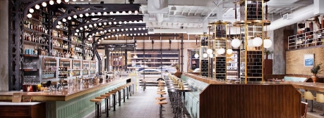 Modern restaurant interior with long wooden bar, high stools, hanging globe lights, and floor-to-ceiling shelves filled with bottles. Large windows in the background show a street view.