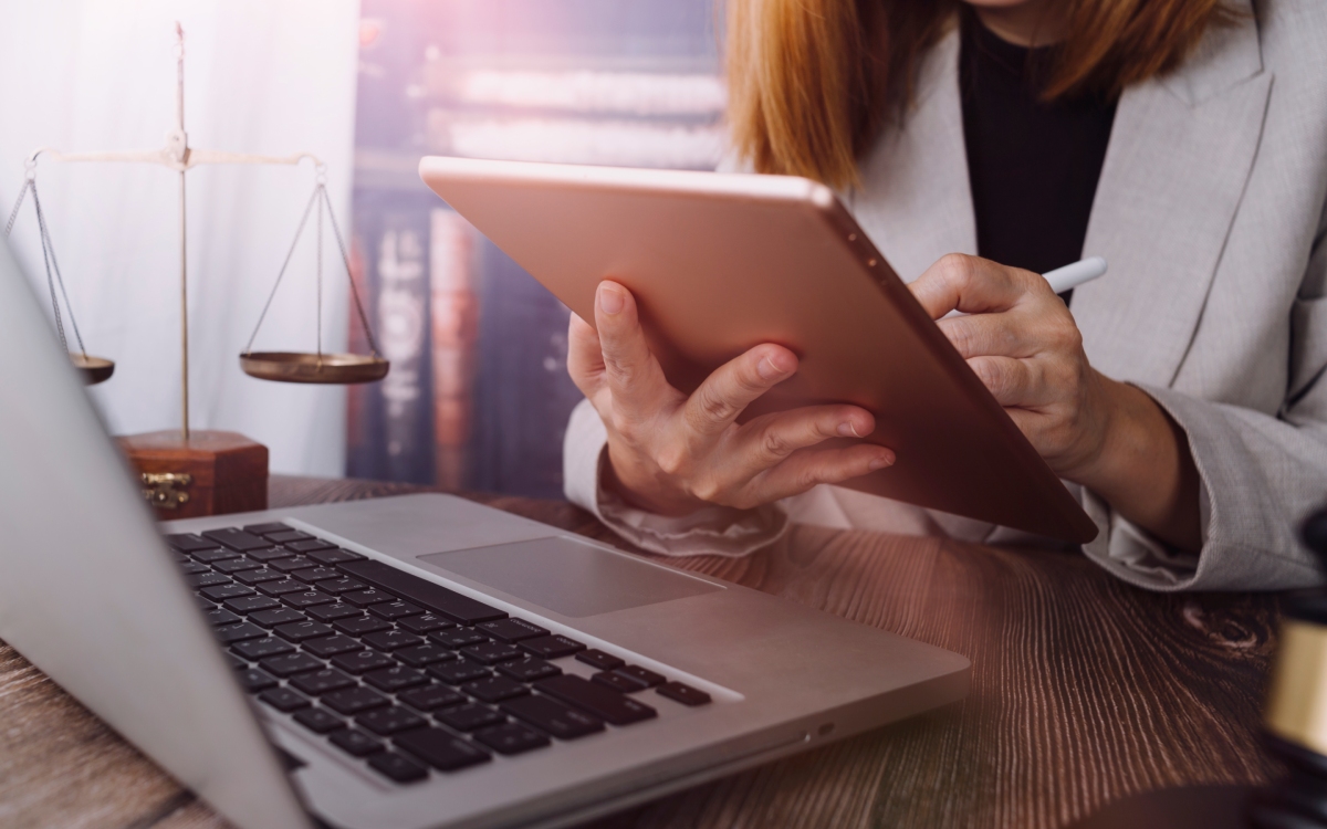Person in business attire using a tablet and stylus at a desk with a laptop, law books, and a justice scale in the background, demonstrating how technology ensures accurate records for modern litigation.