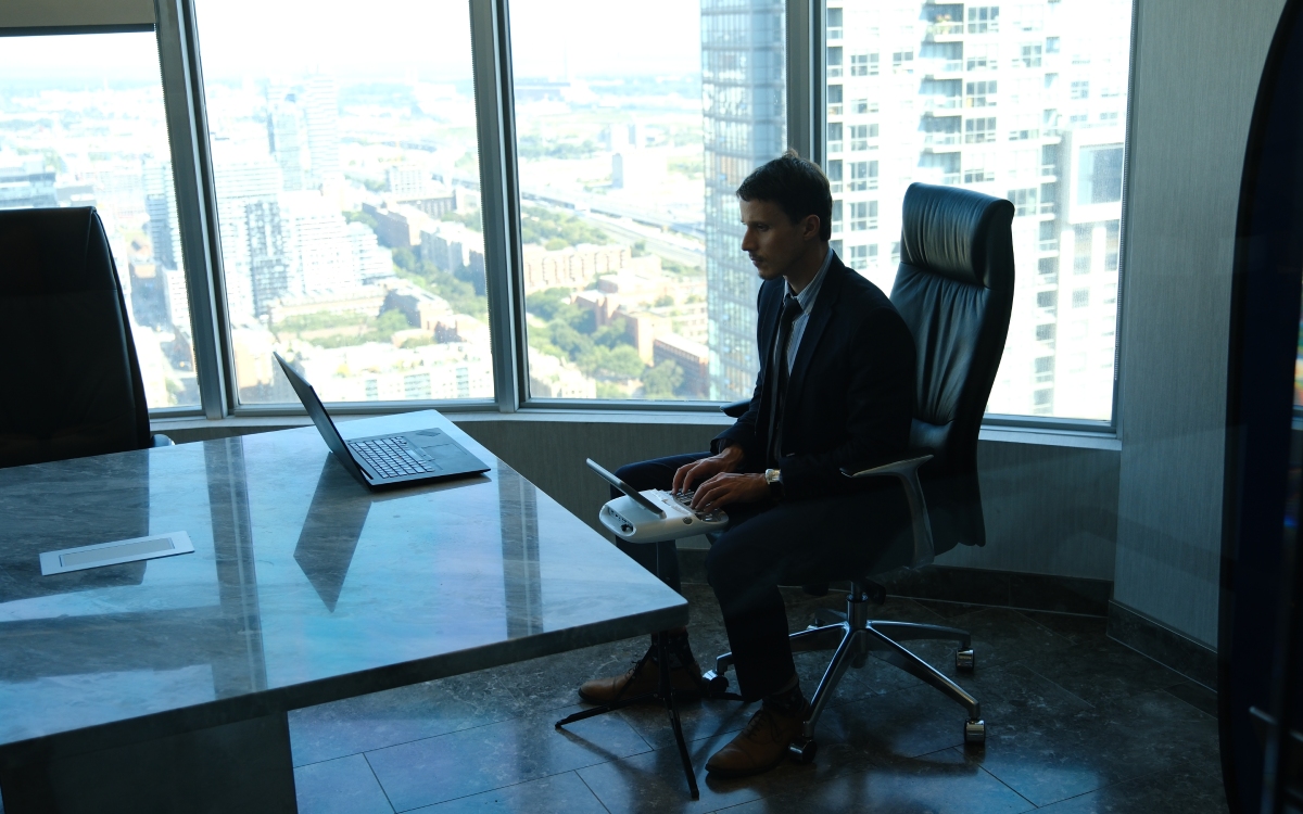 A man sits in an office chair at a desk with a laptop, in front of large windows overlooking a cityscape.