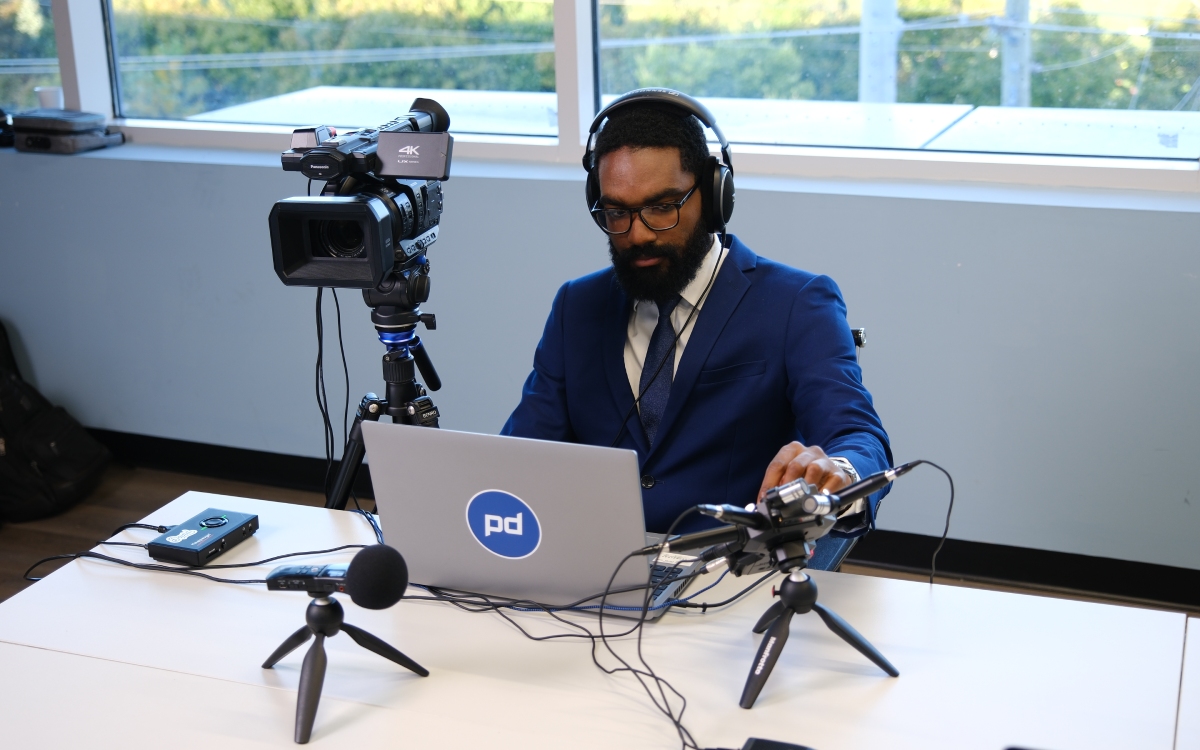 A man in a blue suit operates audio and video equipment at a desk, using a laptop, camera, microphones, and headphones in a bright room with large windows.