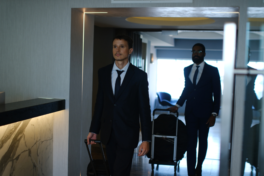 Two men in business suits walk through a hallway pulling suitcases.