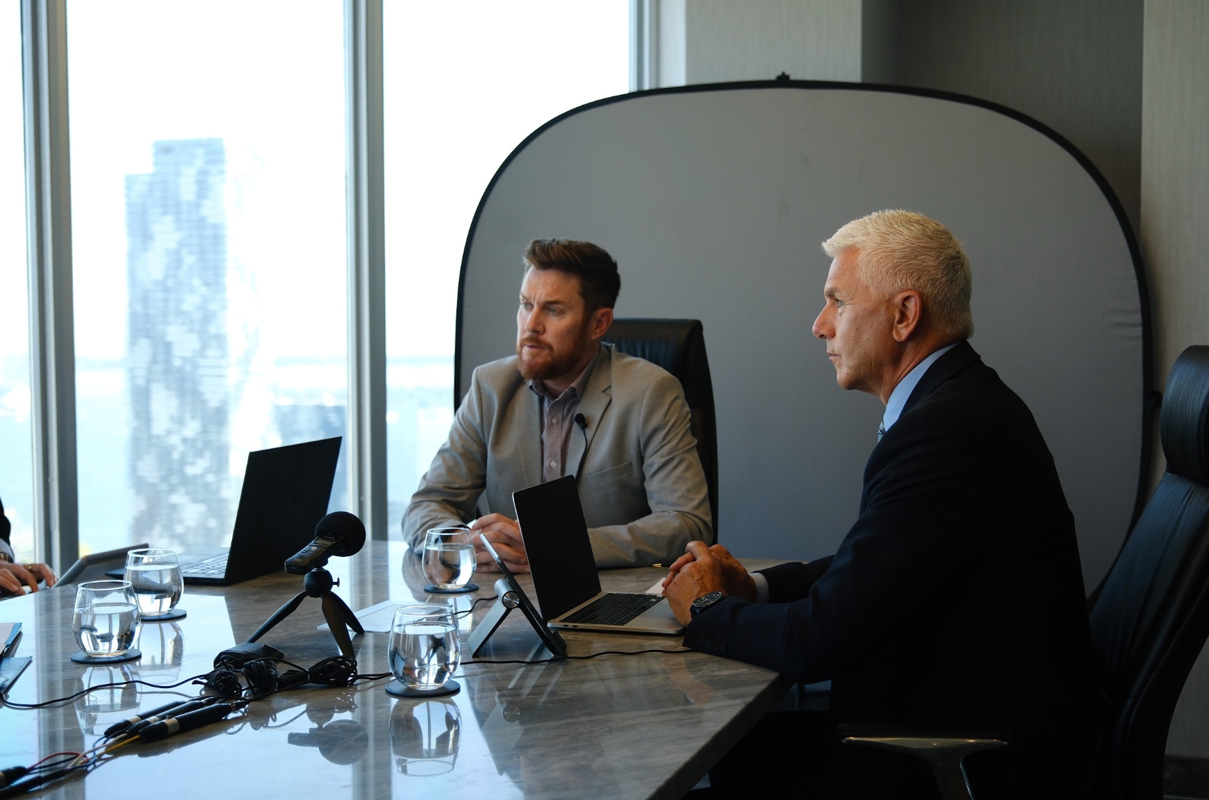 Two men in suits sit at a conference table with laptops, microphones, and water glasses, participating in a meeting with a city view in the background.