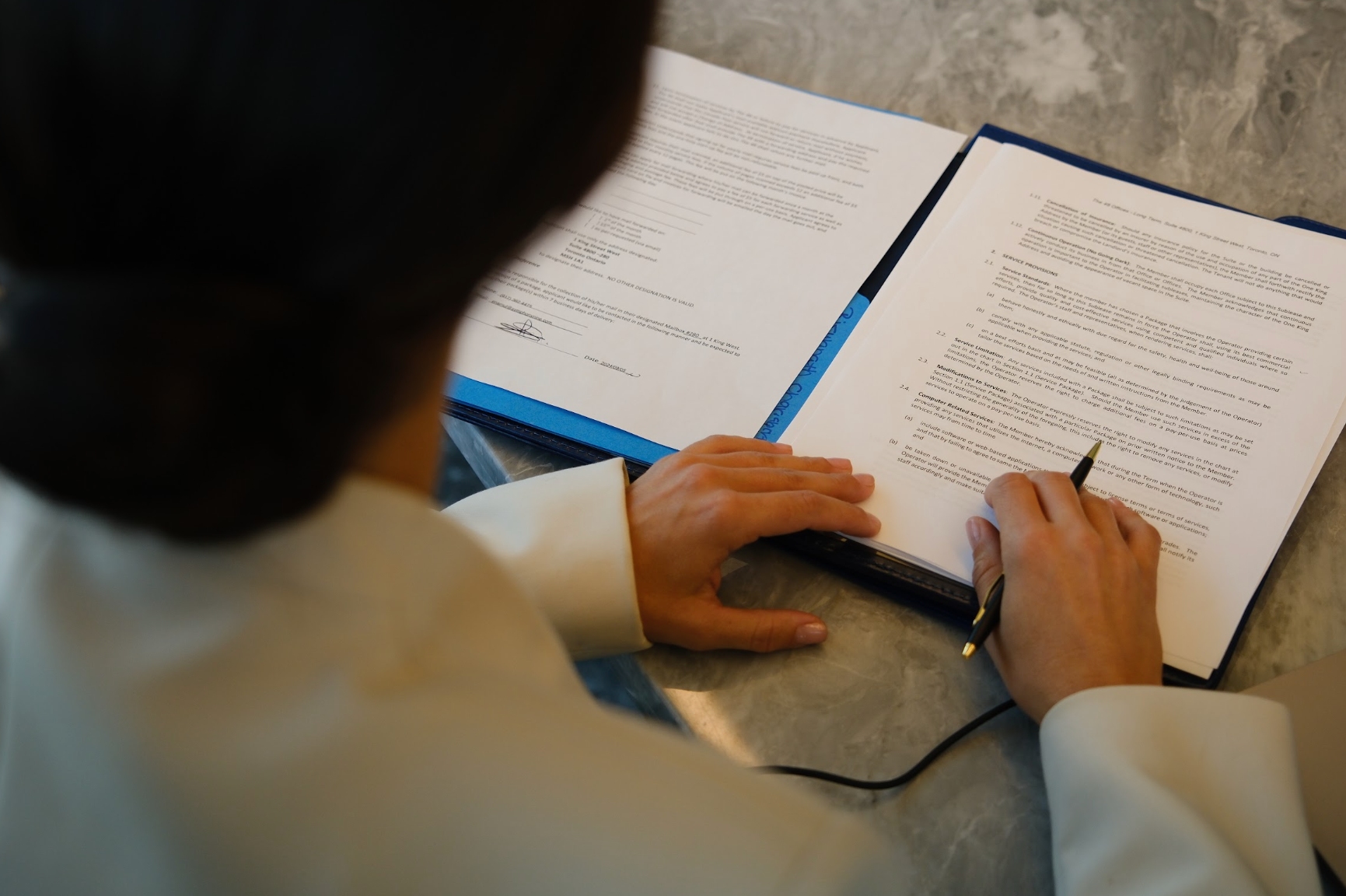 A person in a light-colored blazer reviews and marks documents on a desk, holding a pen and touching printed pages clipped inside a folder.