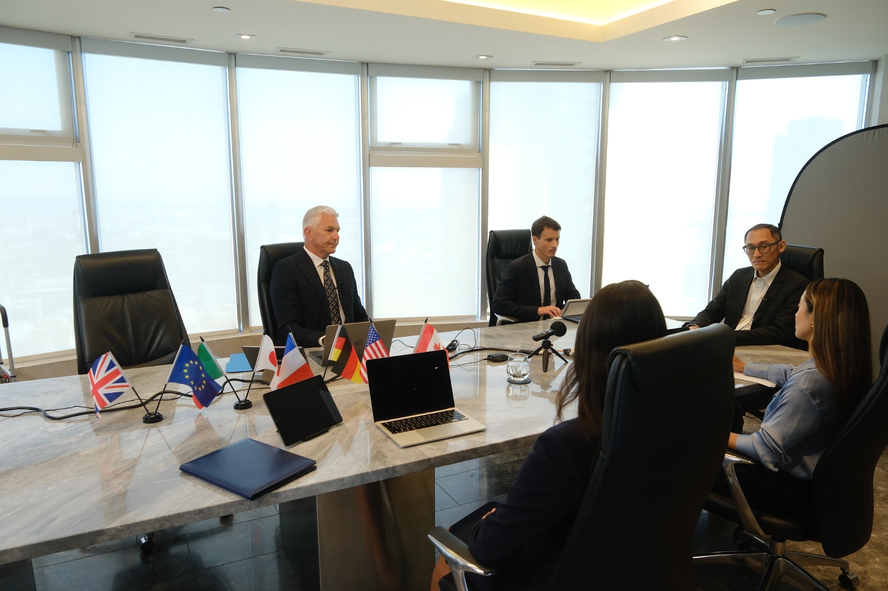 Six people in business attire sit around a conference table with laptops and several international flags, in a modern office with large windows.