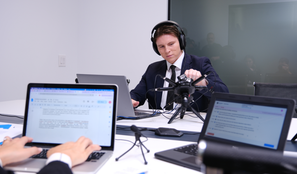 A person in a suit and headphones adjusts a microphone in a conference room. Two laptops with documents open are visible in the foreground.